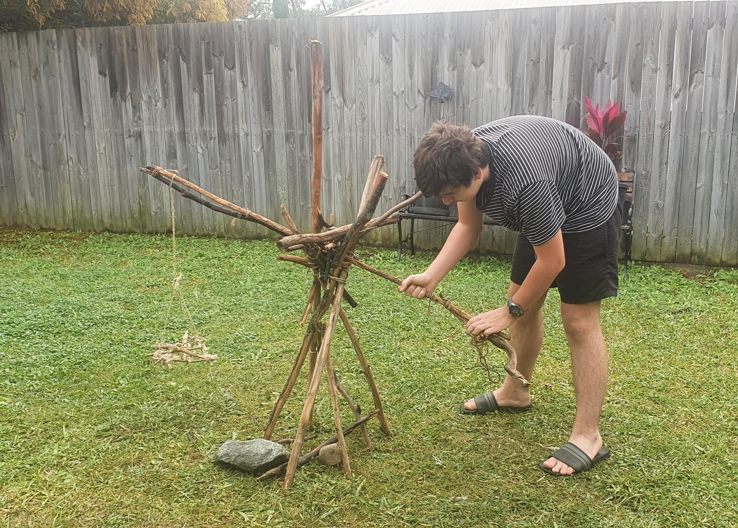 A young man building a stick-and-twine crane structure in a sunny backyard